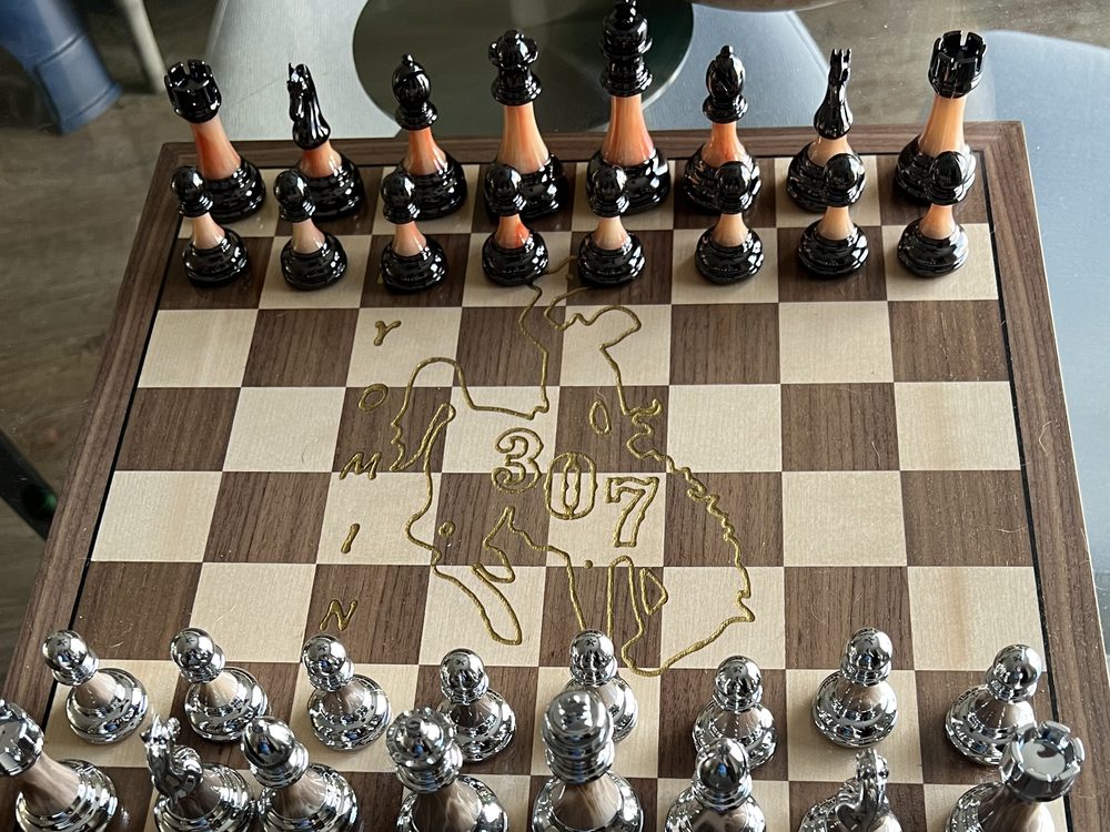 A hardwood chess board with the Wyoming state outline engraved in the centre, chrome and black pieces set up for a game