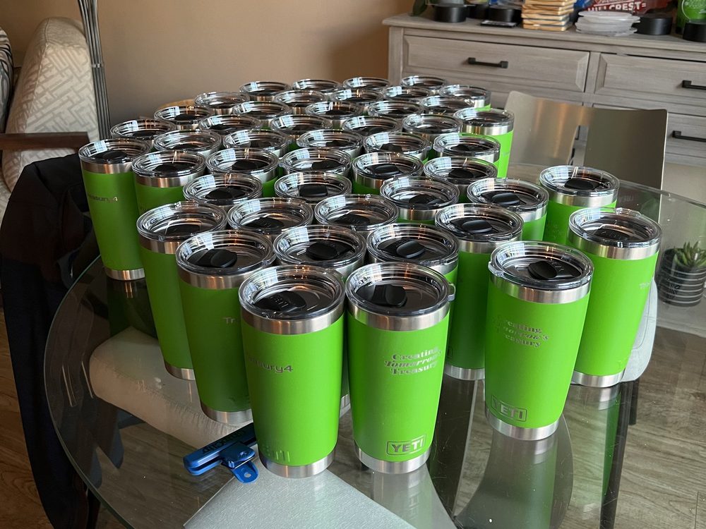About forty green insulated tumblers laser-engraved with a client logo, laid out together after a production run