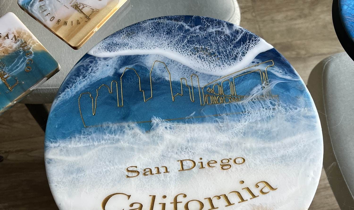 A round hand-poured ocean resin piece with the San Diego skyline engraved in gold beneath white resin foam