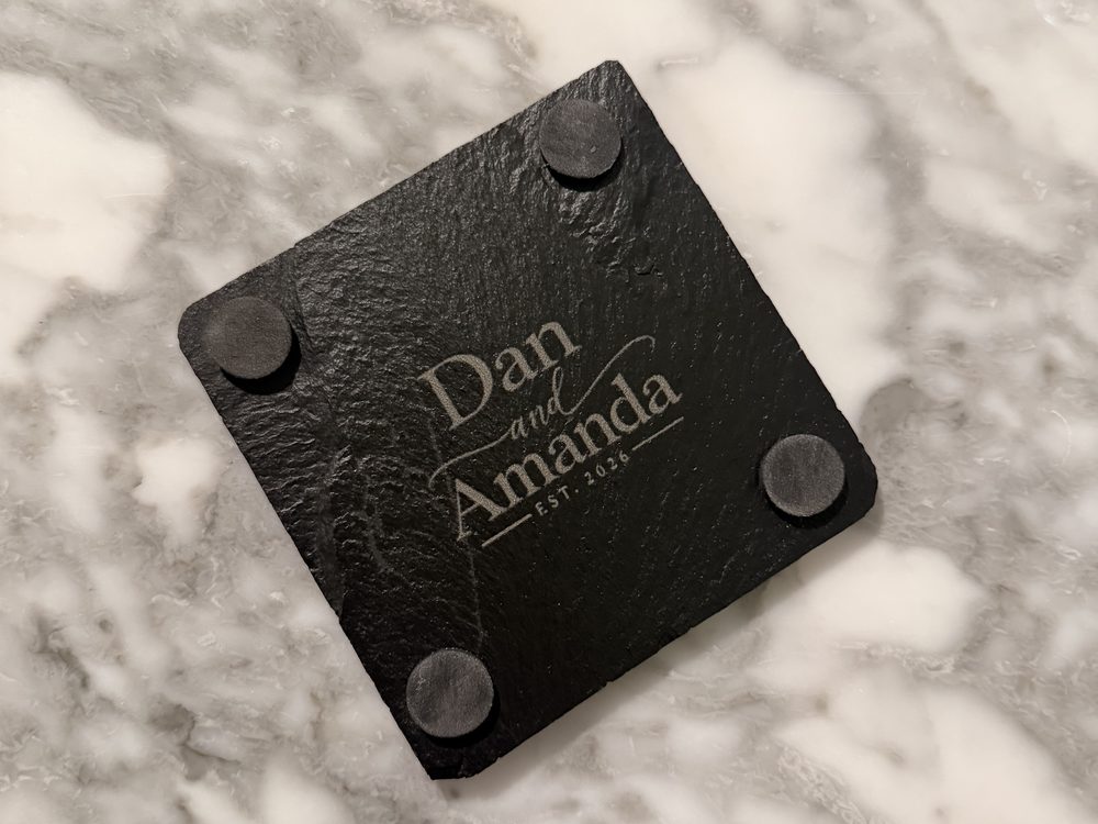 A slate coaster engraved with the names Dan and Amanda and Est. 2026, rubber feet at each corner, on a marble counter