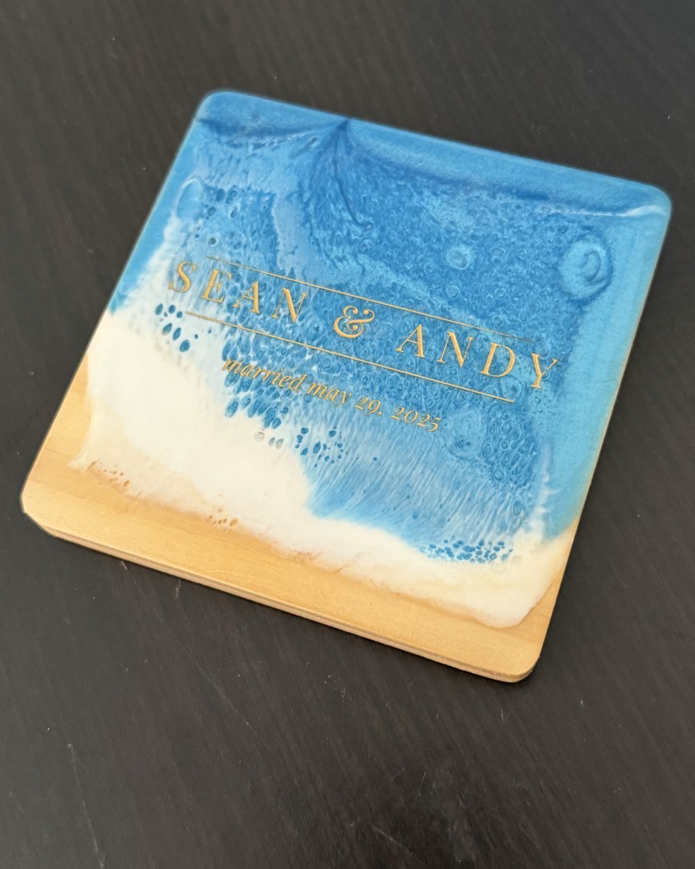 A square wood and ocean resin coaster engraved in gold with Sean and Andy and their wedding date, on a dark table